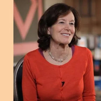 Smiling woman with short dark hair wearing an orange cardigan and layered necklaces sitting indoors.